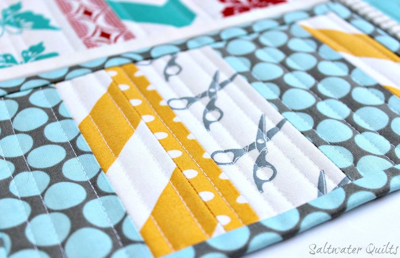 May include: A quilted fabric organizer featuring a patchwork design with light blue and gray polka dots, mustard yellow diagonal stripes, and a white panel with gray scissor icons. The piece is stitched with vertical lines and includes the text Saltwater Quilts.