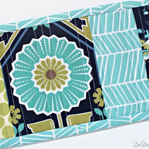 May include: A rectangular quilted fabric mat featuring a central panel with a large teal and olive green flower on a dark navy background, flanked by panels of light green polka dots and a teal and white chevron pattern with floral accents.