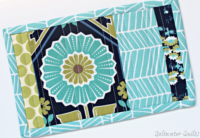May include: A rectangular quilted fabric mat featuring a central panel with a large teal and olive green flower on a dark navy background, flanked by panels of light green polka dots and a teal and white chevron pattern with floral accents.