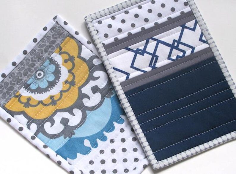 May include: Two rectangular quilted fabric pot holders. One features a yellow, blue, and gray floral pattern with a white and gray polka dot border. The other displays a navy blue section, a white and blue geometric pattern, and a gray polka dot border.