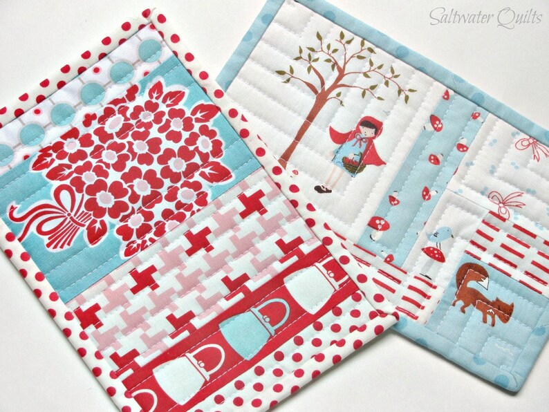 May include: Two rectangular quilted fabric mats featuring patchwork designs. One mat displays a red floral bouquet, red and white geometric patterns, and red polka dots. The other mat depicts a character in a red hood, a tree, and a fox.