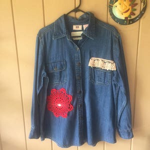 May include: A blue denim long-sleeve button-down shirt featuring a bright red circular crochet doily appliqué on the lower right side and a strip of cream-colored floral lace trim attached to the top of the left chest pocket.