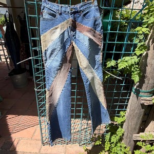 May include: Blue denim jeans with a patchwork design featuring beige, brown, and light blue chevron stripes. The jeans are hanging on a black hanger against a green metal grid.