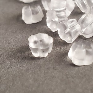 May include: A collection of clear, translucent earring backs scattered on a gray surface. The earring backs are small, with a flower-like shape and a hole in the center. They appear to be made of a soft, flexible material.
