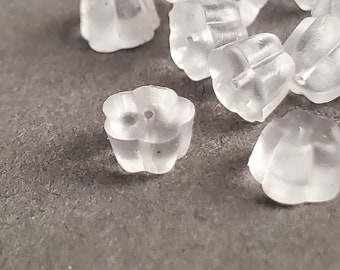 May include: A collection of clear, translucent earring backs scattered on a gray surface. The earring backs are small, with a flower-like shape and a hole in the center. They appear to be made of a soft, flexible material.