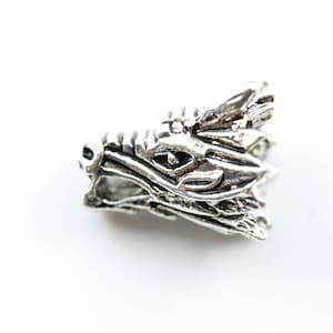 May include: Silver dragon head charm with intricate details. The dragon's head is facing to the right and has a detailed eye and scales.