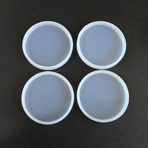 May include: Four clear, round silicone coaster molds with white rims. The molds are arranged in a square formation against a dark background. These molds are designed for creating custom coasters.