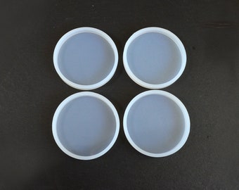 May include: Four clear, round silicone coaster molds with white rims. The molds are arranged in a square formation against a dark background. These molds are designed for creating custom coasters.