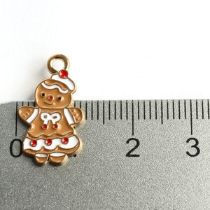 Może przedstawiać: Złoty charms w kształcie piernikowego ludzika z białą polewą i czerwonymi akcentami. Charms ma około 2,5 cm wysokości i jest pokazany obok linijki.