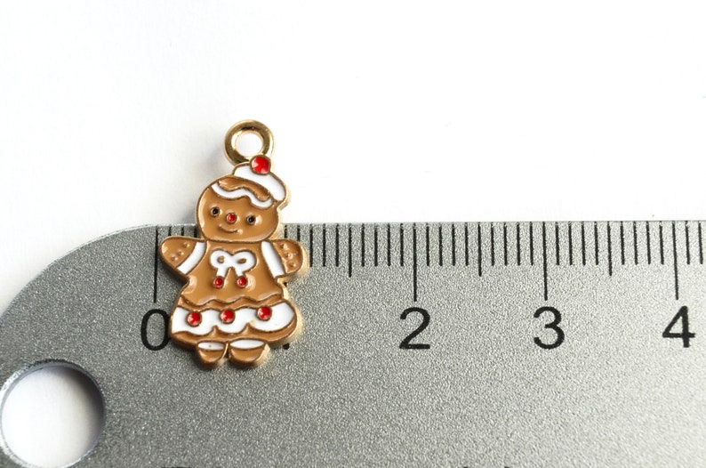 Może przedstawiać: Złoty charms w kształcie piernikowego ludzika z białą polewą i czerwonymi akcentami. Charms ma około 2,5 cm wysokości i jest pokazany obok linijki.