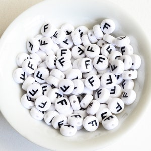 May include: A collection of white, round beads with the letter "F" in black, displayed in a white bowl. The beads are evenly sized and have a flat surface with a hole for stringing. Ideal for jewelry making and crafts.