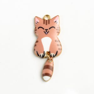 May include: A gold charm with a cartoon image of a brown and white striped cat with a pink nose and a white belly. The cat is sitting with its tail dangling down. The tail is also brown and white striped.