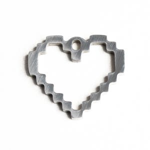 May include: A silver metal heart-shaped charm with a pixelated design. The charm has a hole at the top for attaching to a necklace or bracelet.