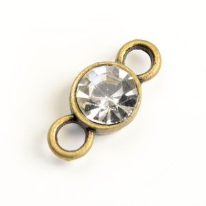 May include: Antique gold-tone connector with a clear crystal rhinestone in the center. The connector has two loops on either side for attaching jewelry.