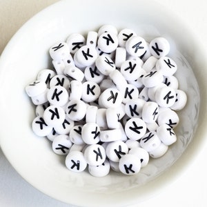 May include: A white bowl filled with white round beads with the letter "K" printed in black on each bead.