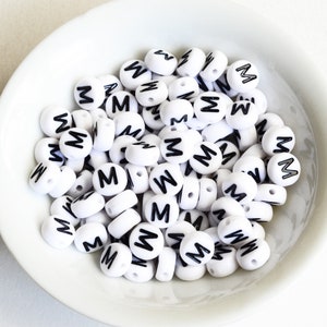 May include: A white bowl filled with white plastic letter beads, each bead has a black letter "M" printed on it.