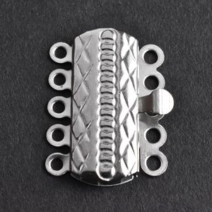May include: A silver-colored multi-strand jewelry clasp with a textured, diamond-patterned surface. The clasp has multiple loops for attaching strands and a secure closure mechanism. The background is a dark gray.
