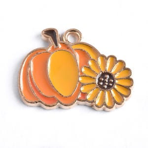 May include: A gold-toned charm featuring a pumpkin and a sunflower. The pumpkin is orange and yellow with a brown stem. The sunflower has yellow petals and a brown center. The charm has a loop for attaching to jewelry or other crafts.