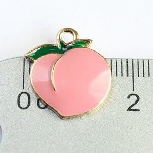 May include: Gold-tone peach charm with a green leaf, measuring approximately 1 inch in diameter. The charm is pink and has a shiny finish.