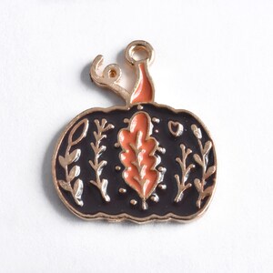 May include: A black pumpkin charm with gold trim and a gold stem. The pumpkin features an orange leaf design in the center and floral accents. A small heart is visible on the right side of the pumpkin.