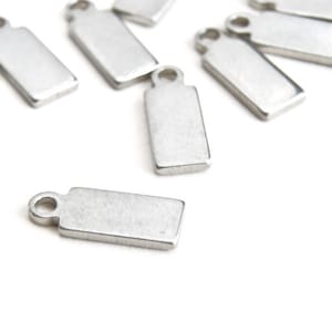 May include: A collection of small, rectangular silver metal charms with a loop at the top. The charms are scattered on a white surface, with some in focus and others blurred. The charms have a brushed metal finish.