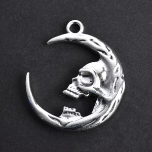 May include: Silver pendant charm in the shape of a crescent moon with a skull design. The skull is detailed with eye sockets and a nose.