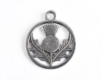 May include: A silver-toned pendant featuring a detailed thistle design within a circular frame. The thistle, a symbol of Scotland, is depicted with textured petals and leaves. The pendant has a loop at the top for attachment.