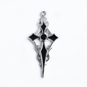 May include: A silver-toned pendant in the shape of a cross, featuring black enamel accents and a central black stone. The design includes ornate, stylized details and a loop for hanging. The pendant is set against a white background.