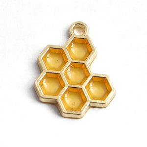 May include: Gold-toned metal charm shaped like a honeycomb with a translucent yellow resin fill. The charm has a loop at the top for attaching to jewelry or other crafts.