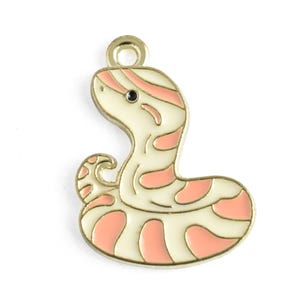 May include: A gold-toned metal charm shaped like a coiled snake with cream-colored enamel fill and soft pink striped patterns. The snake has a single black dot for an eye and a small circular loop at the top for attachment to jewelry or accessories.