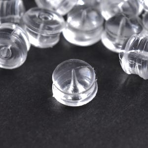 May include: A collection of clear, transparent earring backs. The earring backs are made of a soft, flexible plastic material. The earring backs are round with a small post hole in the center. The earring backs are scattered on a dark surface.