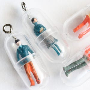 May include: Miniature figurines inside clear capsule pendants. The figurines are painted with various colors, including blue, orange, and green. Each pendant has a silver-colored loop at the top for hanging. The pendants are on a white background.
