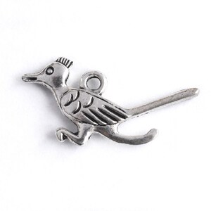 May include: A silver-colored metal charm in the shape of a roadrunner bird. The charm has a loop at the top for attaching to a necklace or bracelet. The bird is depicted in a running pose with its tail extended.