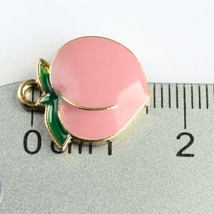 May include: A gold-colored peach charm with a green leaf, measuring approximately 1 inch in length. The charm is pink and has a shiny finish.