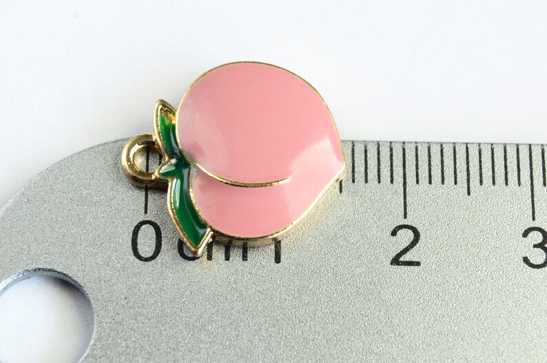 May include: A gold-colored peach charm with a green leaf, measuring approximately 1 inch in length. The charm is pink and has a shiny finish.