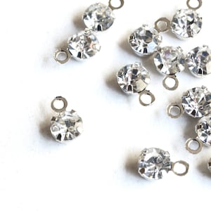 May include: Scattered on a white surface are several clear, round crystal charms with silver-colored loops. The crystals sparkle, reflecting light. These charms are likely used for jewelry making or other craft projects.