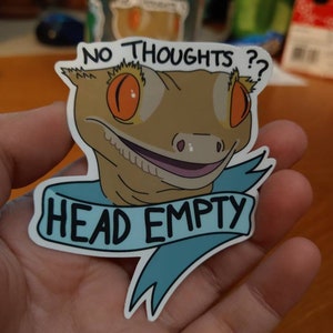 Può includere: Un adesivo raffigurante un geco dei cartoni animati con occhi arancioni e un'espressione sorridente. Il testo "NO THOUGHTS ??" è sopra la testa del geco e uno striscione sottostante recita "HEAD EMPTY" in un carattere giocoso. L'adesivo ha un bordo bianco.