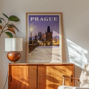 a framed poster of Prague, Czech Republic, with a snowy cityscape in the background, hanging on a wall next to a wooden cabinet and a lamp.