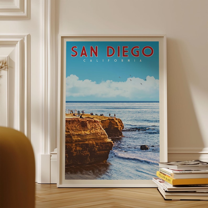 A framed poster of a scenic coastal landscape with cliffs and people enjoying the view, displayed on a wall next to a stack of books.