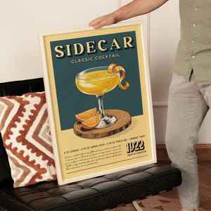 A framed poster of a classic cocktail called "Sidecar" is being held by a person in a living room.