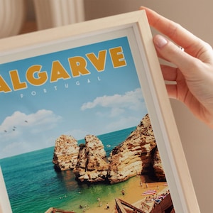 A person's hand is holding a framed picture of a scenic coastal landscape in Portugal, featuring rocky cliffs, a beach, and a clear blue sky.