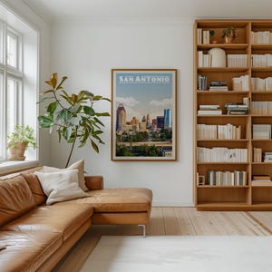 a living room with a large window, a brown leather sofa, a bookshelf filled with books, and a framed poster of San Antonio, Texas.