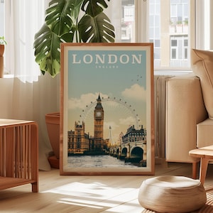 A framed poster of London, England, featuring the iconic Big Ben and the London Eye, is displayed in a living room setting with a potted plant and a window in the background.