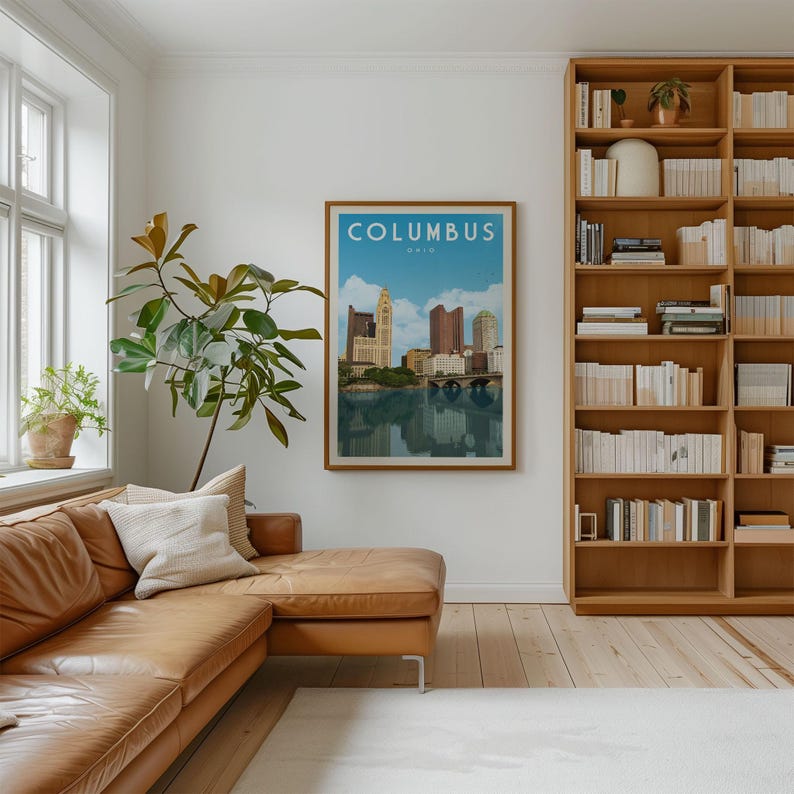 a cozy living room with a large window, a comfortable leather couch, a bookshelf filled with books, and a framed poster of the city of Columbus hanging on the wall.