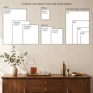 a wall with a variety of framed artworks in different sizes, ranging from small to large, and international paper sizes, such as A1, A3, and A4. The artworks are arranged on a wooden cabinet or dresser, which also has a vase, a cocktail shaker, and some glasses and fruit on it.