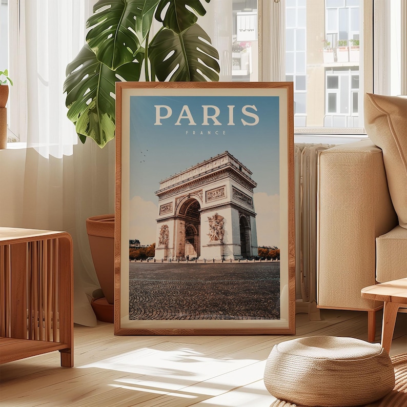A framed poster of the Arc de Triomphe in Paris, France, is displayed in a living room setting with a large window and a potted plant in the background.