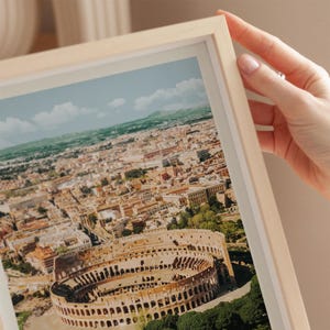 A hand is holding a framed photograph of the Colosseum in Rome, Italy, with the city's skyline visible in the background.