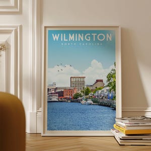 A framed poster of a cityscape with a river in the foreground, displaying the text "WILMINGTON NORTH CAROLINA" and a few boats on the water.