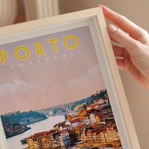 A person's hand is holding a framed poster of the city of Porto, Portugal, with a colorful and detailed view of the city's skyline and river.
