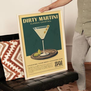 A framed poster of a Dirty Martini cocktail is being held by a person, with a vintage-style design featuring a martini glass and garnishes.